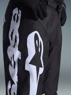 Pantaloni Alpinestars Racer Air Riway 2026, pantaloni da motocross neri con grande logo Alpinestars bianco sulla coscia, primo piano del tessuto tecnico e della chiusura regolabile in vita, indossati con guanti neri per la guida fuoristrada