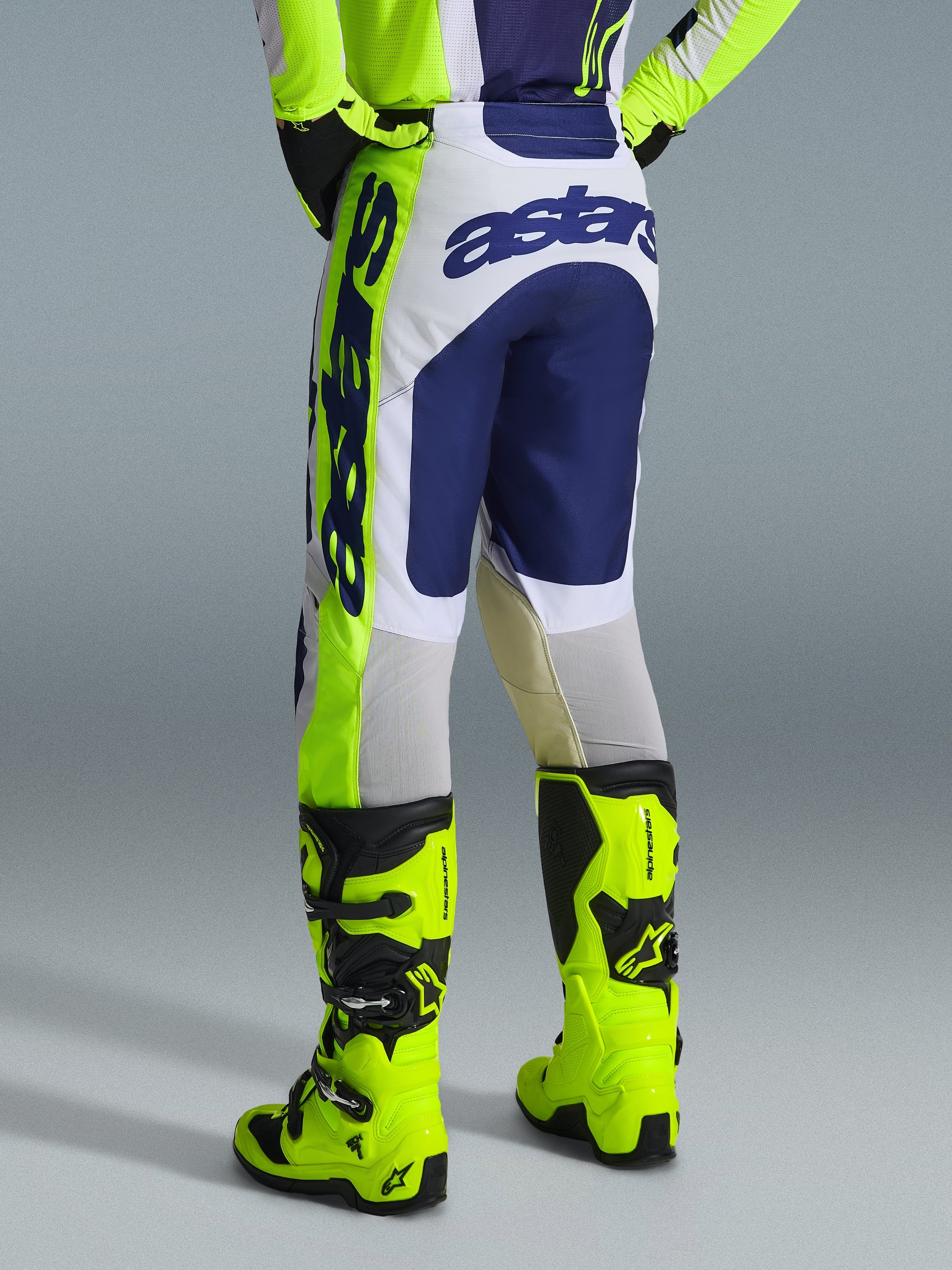 Alpinestars 2026 Racer Air Riway Pants, pantaloni da motocross, grigio chiaro giallo fluo e blu, vista posteriore con marchio blu in grassetto sulla seduta, indossati con maglia coordinata e stivali giallo fluo brillanti, design ergonomico race-fit