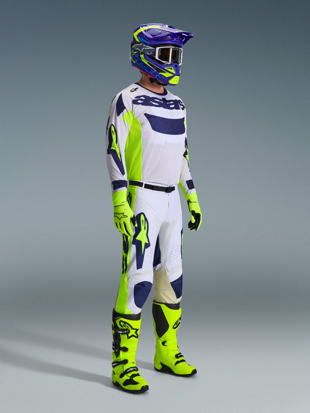 Pilota di motocross con equipaggiamento completo da gara, pantaloni 2026 Racer Air Riway in grigio chiaro, giallo fluo e blu, maglia coordinata bianca e blu, guanti e stivali giallo fluo, casco blu e giallo, outfit coordinato per prestazioni professionali off-road