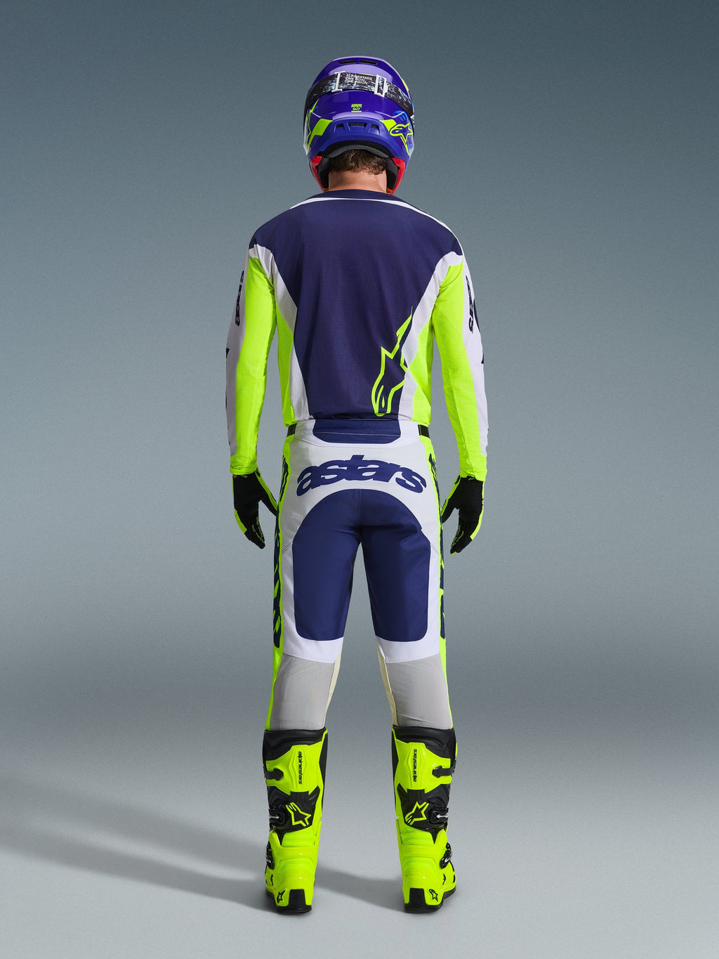 Pilota di motocross visto da dietro che indossa i pantaloni Alpinestars 2026 Racer Air Riway in grigio chiaro, giallo fluo e blu, abbinati a una maglia coordinata e stivali giallo brillante, per un set completo da gara off-road professionale.