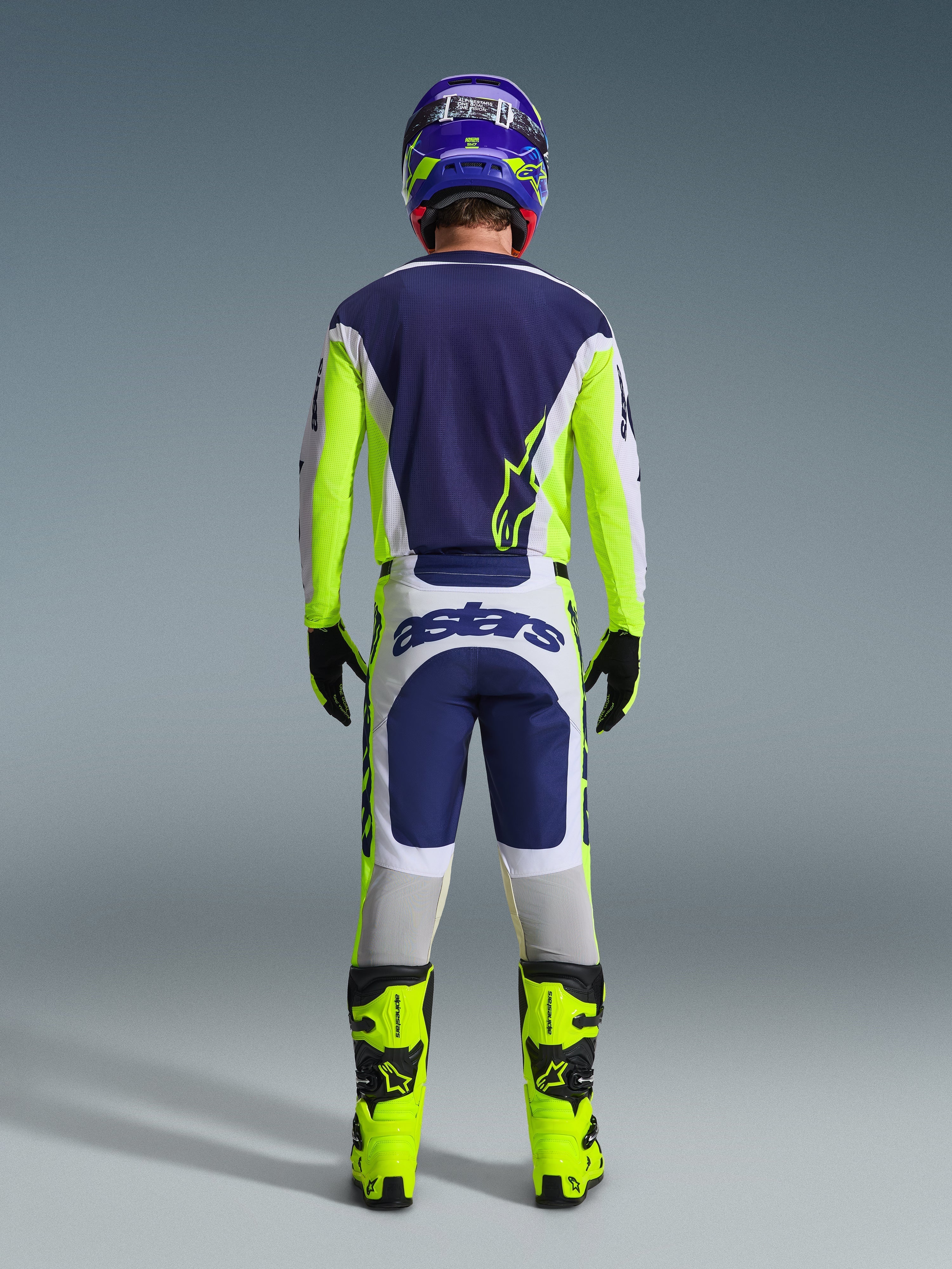 Pilota di motocross visto da dietro che indossa i pantaloni Alpinestars 2026 Racer Air Riway in grigio chiaro, giallo fluo e blu, abbinati a una maglia coordinata e stivali giallo brillante, per un set completo da gara off-road professionale.