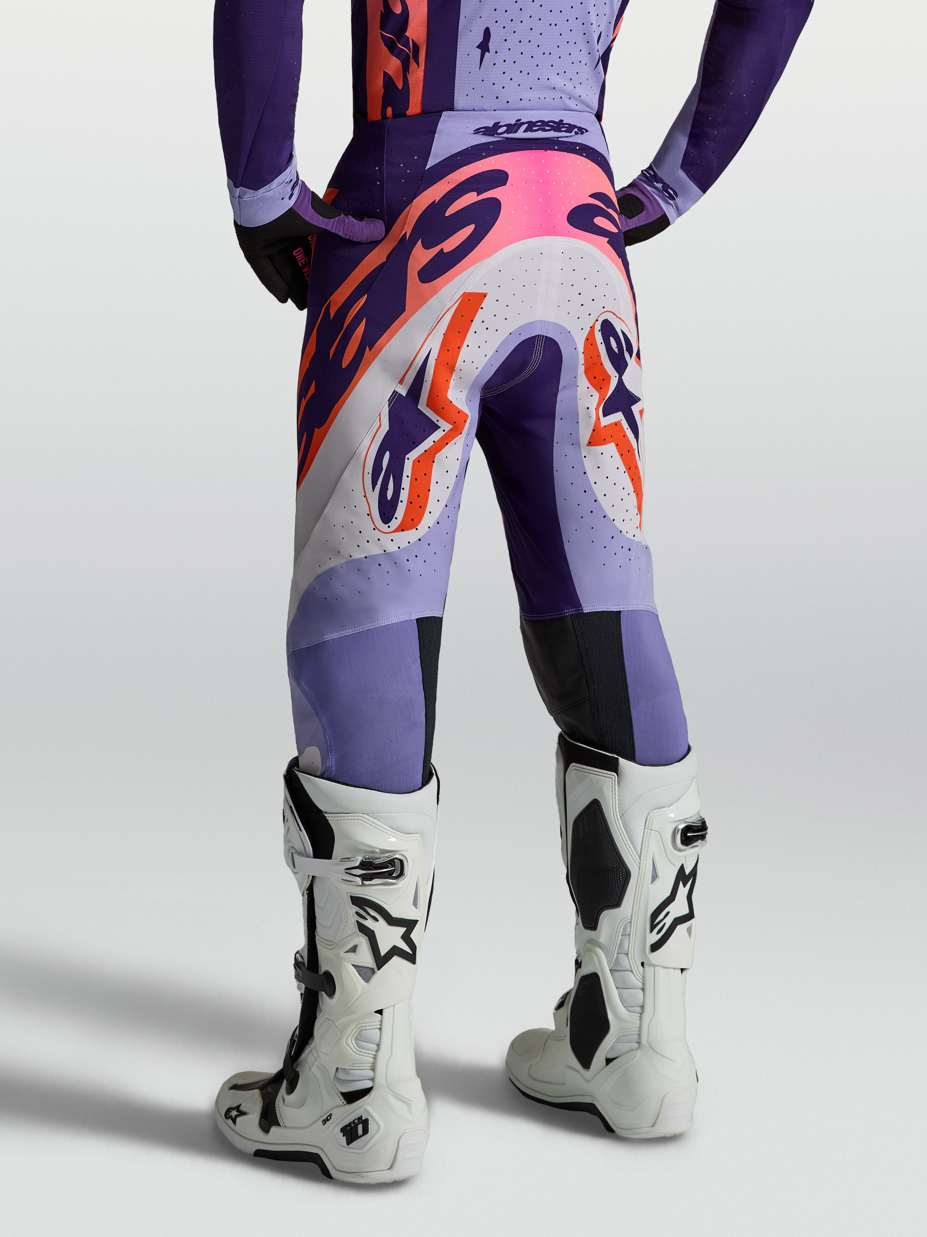Alpinestars Pantaloni Supertech Dyna, pantaloni da motocross per uomo, viola e rosa fluo, in tessuto ripstop elastico e traspirante, vestibilità atletica, vista posteriore indossati con stivali bianchi.