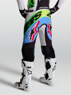 Alpinestars Pantaloni Supertech Dyna, pantaloni motocross da uomo, colore lilla chiaro, verde fluo e nero con rinforzi in pelle, indossati con stivali bianchi, abbigliamento racing off-road