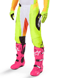 Alpinestars 2025 Techstar Melt Pants, pantaloni da motocross, giallo neon con dettagli bianchi e arancioni, motivi laterali neri, indossati con stivali da motocross rosa acceso e guanti giallo neon, con perforazioni per la ventilazione, abbigliamento tecnico resistente per il fuoristrada