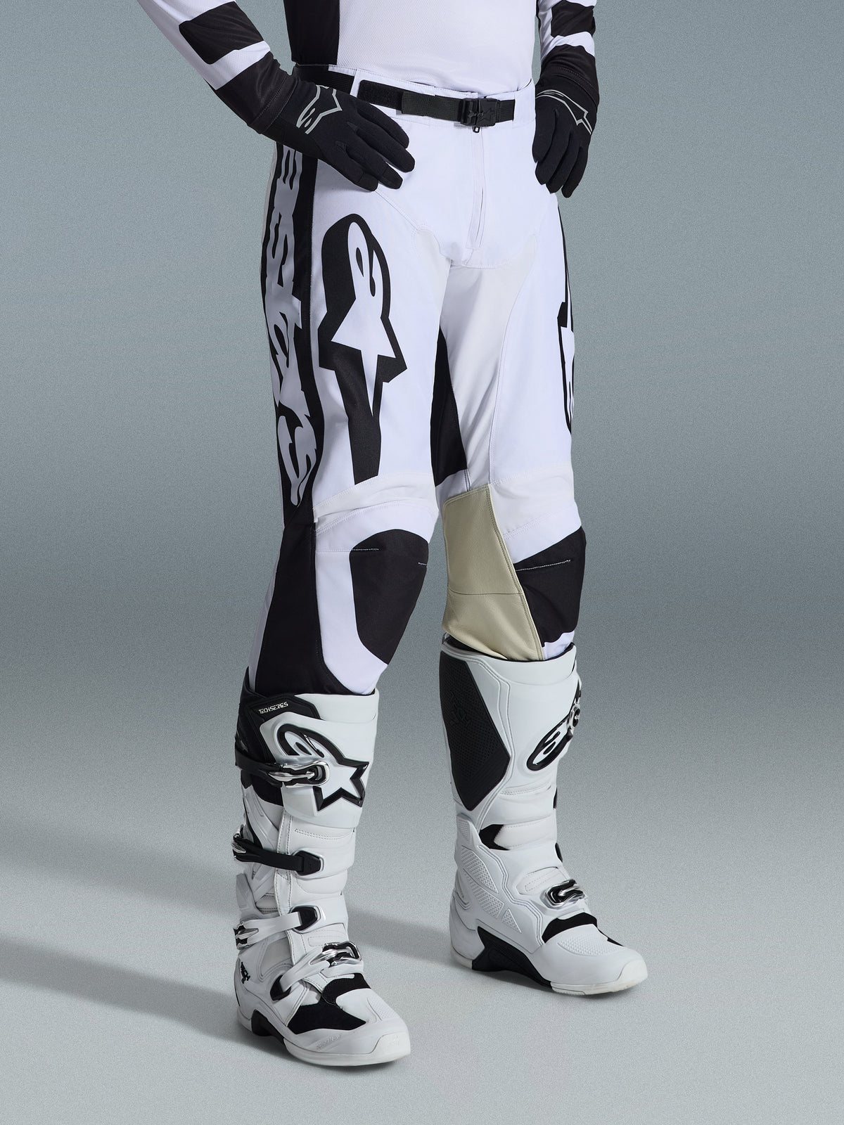 Pantaloni Alpinestars Racer Riway 2026, pantaloni da motocross, bianco e nero con grafiche e loghi Alpinestars neri, rinforzi interni in pelle color cuoio sulle ginocchia, indossati con stivali da motocross bianchi e neri e guanti neri, abbigliamento professionale da gara per il fuoristrada