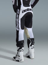 Alpinestars 2026 Racer Riway Pants, pantaloni da motocross, bianco e nero, vista posteriore con branding nero marcato su sedile e fianchi, pannelli interni delle ginocchia in pelle di capra color tan, indossati con stivali da motocross coordinati bianchi e neri.