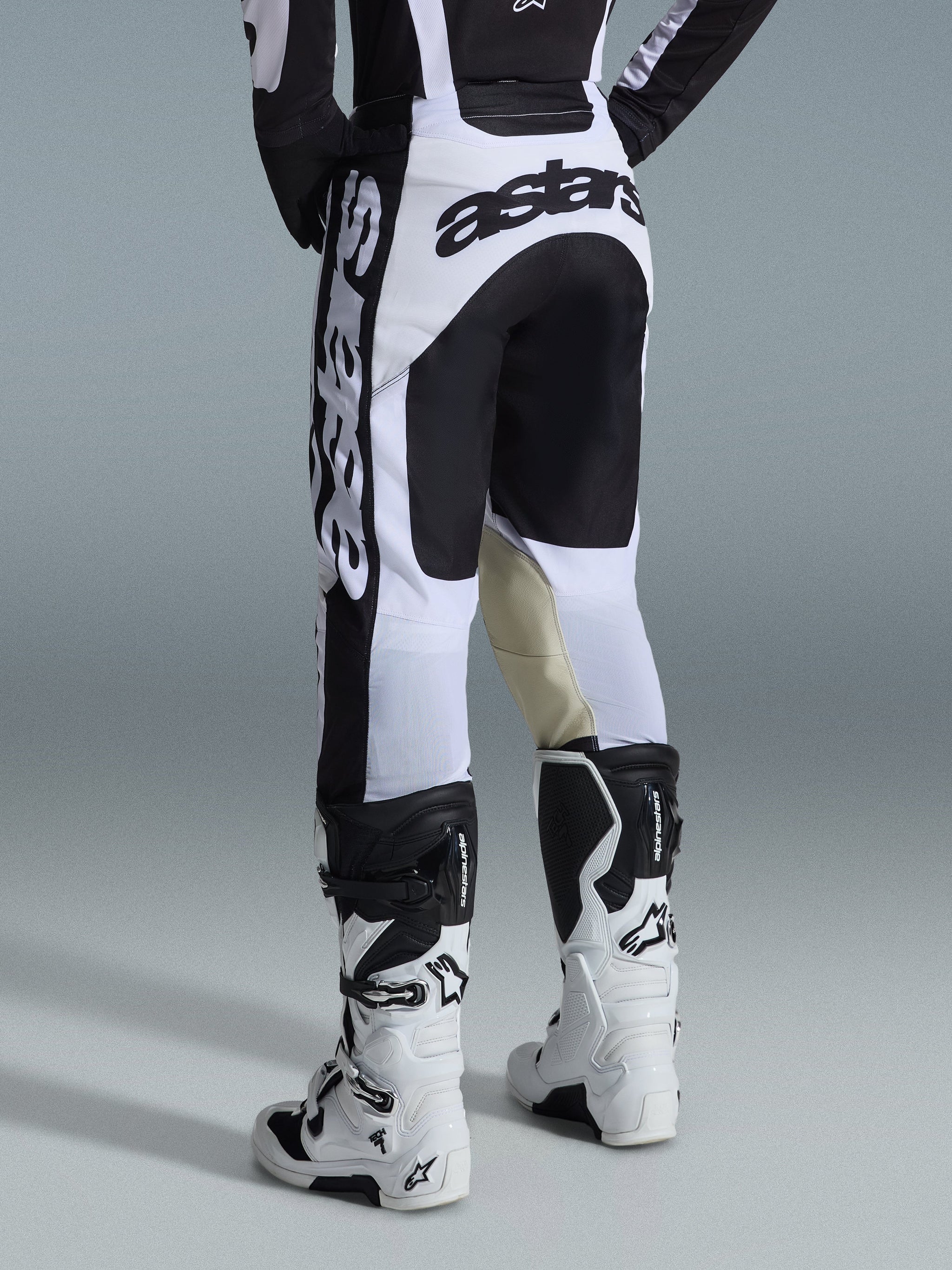 Alpinestars 2026 Racer Riway Pants, pantaloni da motocross, bianco e nero, vista posteriore con branding nero marcato su sedile e fianchi, pannelli interni delle ginocchia in pelle di capra color tan, indossati con stivali da motocross coordinati bianchi e neri.