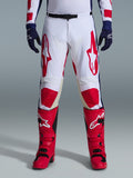 Alpinestars Racer Riway Pants 2026, pantaloni da motocross, bianco, blu e rosso, indossati con stivali rossi e guanti blu navy, con grandi loghi rossi sulle cosce e pannelli rossi sulle ginocchia, design sportivo pre-curvato con fibbia in vita regolabile.
