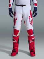 Alpinestars Racer Riway Pants 2026, pantaloni da motocross, bianco, blu e rosso, indossati con stivali rossi e guanti blu navy, con grandi loghi rossi sulle cosce e pannelli rossi sulle ginocchia, design sportivo pre-curvato con fibbia in vita regolabile.