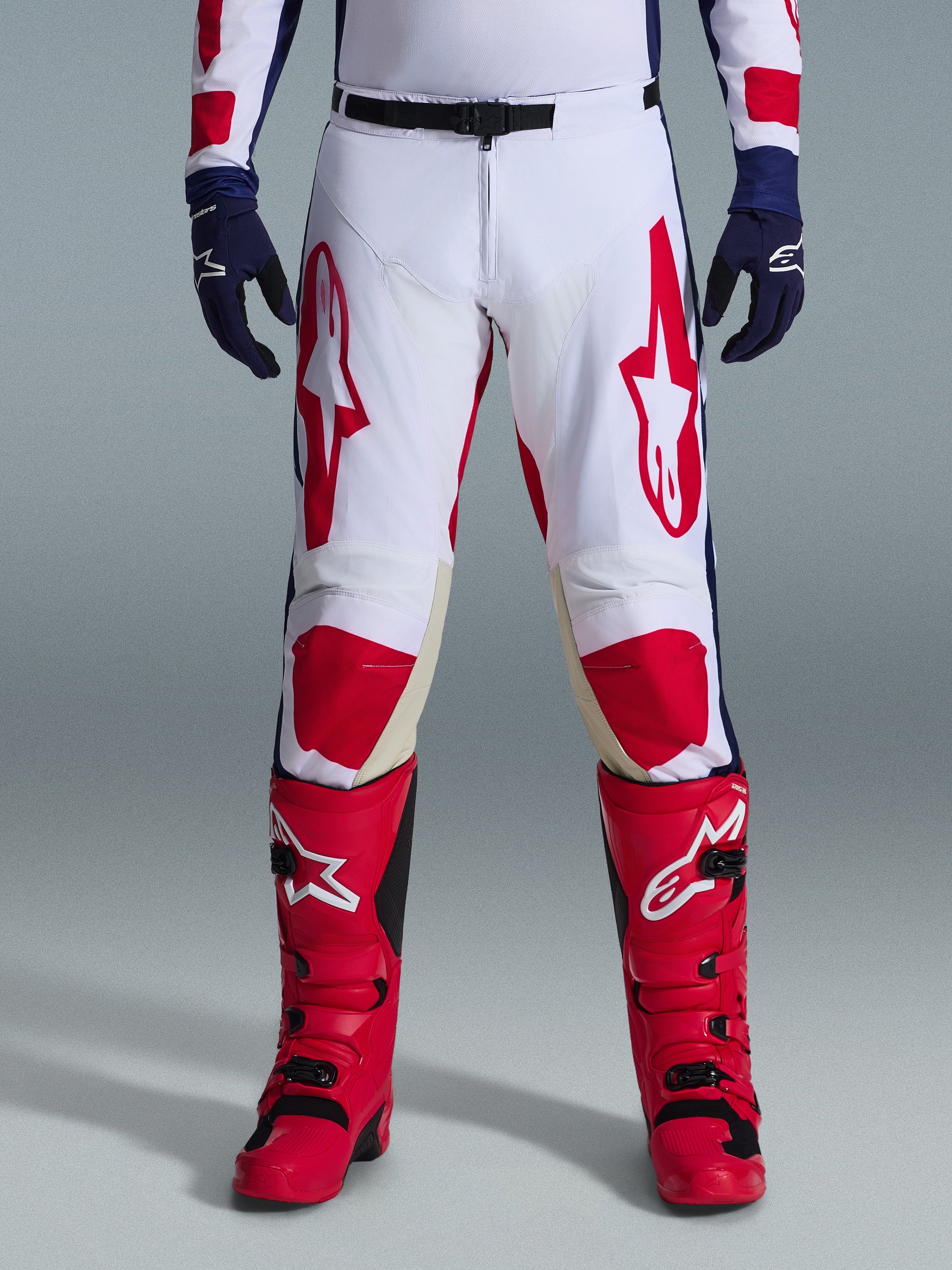 Alpinestars Racer Riway Pants 2026, pantaloni da motocross, bianco, blu e rosso, indossati con stivali rossi e guanti blu navy, con grandi loghi rossi sulle cosce e pannelli rossi sulle ginocchia, design sportivo pre-curvato con fibbia in vita regolabile.