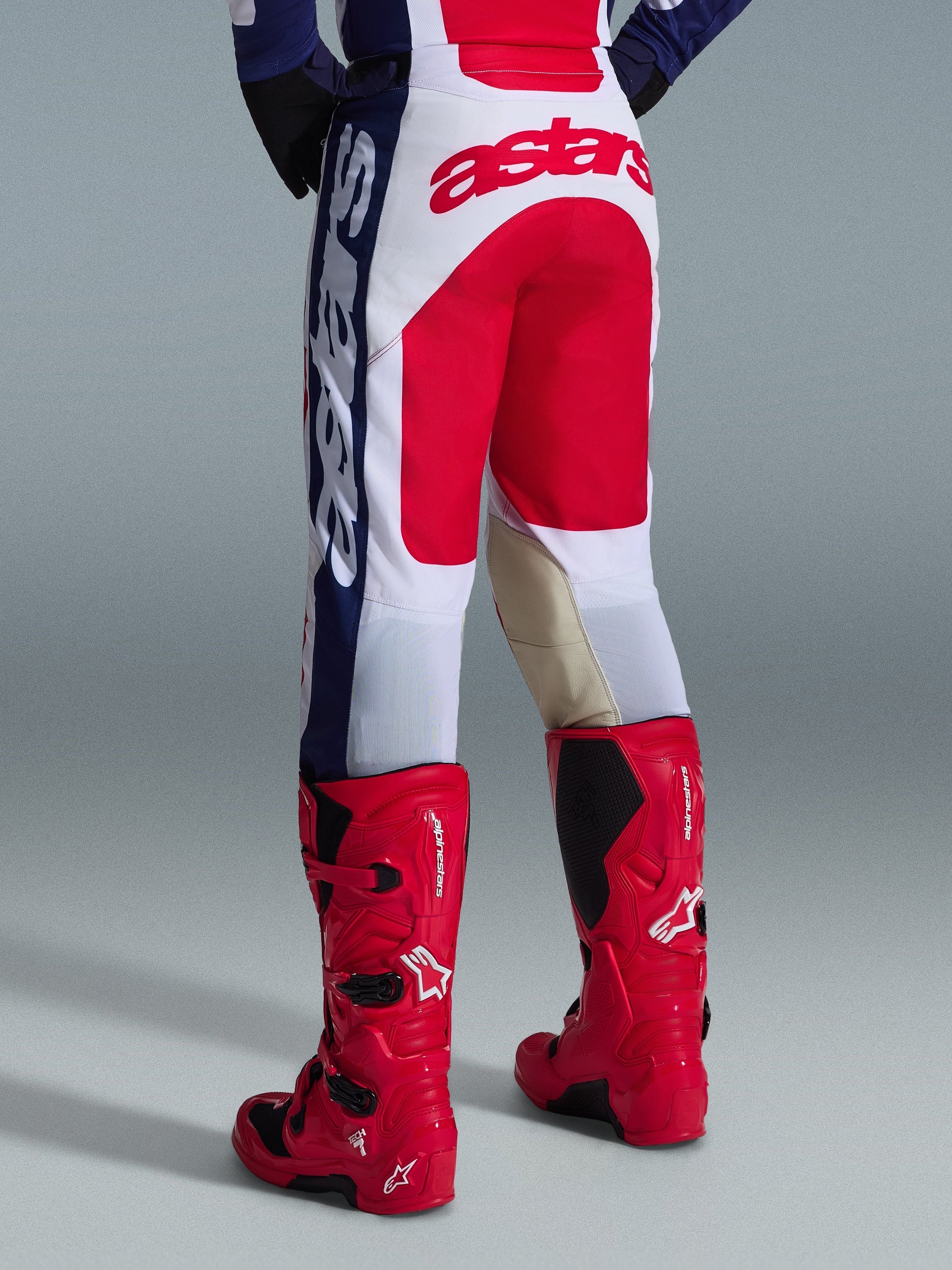 Pantaloni Alpinestars 2026 Racer Riway, pantaloni da motocross, bianco, blu e rosso, vista posteriore con grande logo Astars rosso sulla seduta e pannelli interni alle ginocchia in pelle marrone chiaro, indossati con stivali da motocross Alpinestars rossi, abbigliamento tecnico per gare off-road