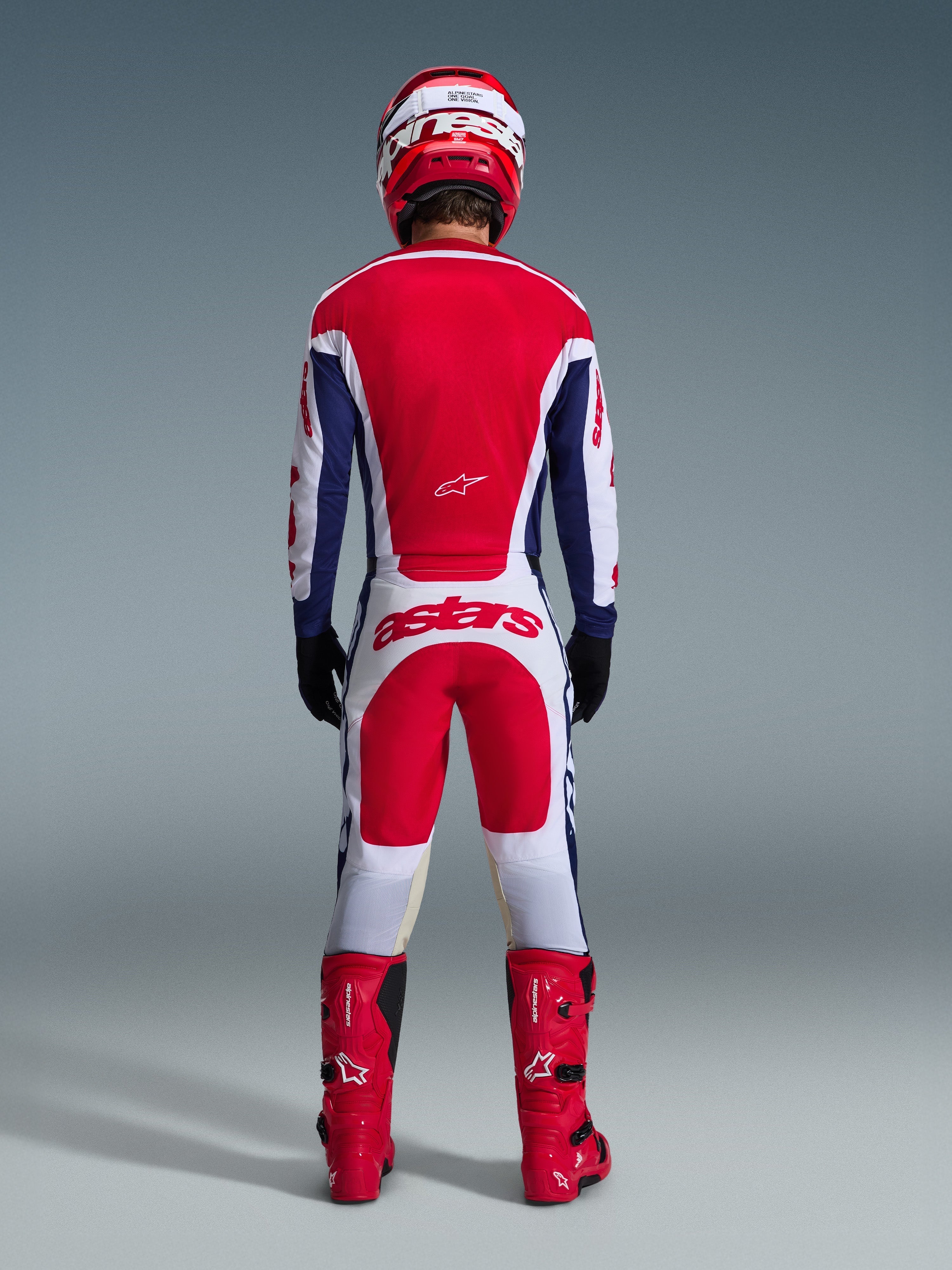 Pilota di motocross visto da dietro con abbigliamento completo da gara, pantaloni Alpinestars Racer Riway 2026 in bianco blu e rosso con maglia abbinata, stivali e casco rossi, grande logo 'astars' sulla seduta, completo da motocross coordinato.