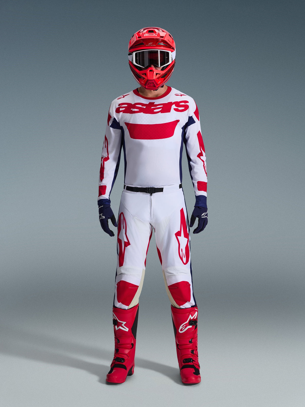 Pilota di motocross che indossa il completo Alpinestars Racer Riway 2026, maglia bianca con marchio "astars" rosso e pannelli laterali blu, pantaloni bianchi con grafiche rosse, casco da gara e stivali rossi, guanti blu scuro, abbigliamento completo coordinato da motocross