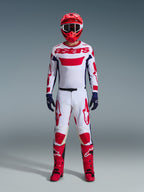 Pilota di motocross che indossa il completo Alpinestars Racer Riway 2026, maglia bianca con marchio "astars" rosso e pannelli laterali blu, pantaloni bianchi con grafiche rosse, casco da gara e stivali rossi, guanti blu scuro, abbigliamento completo coordinato da motocross