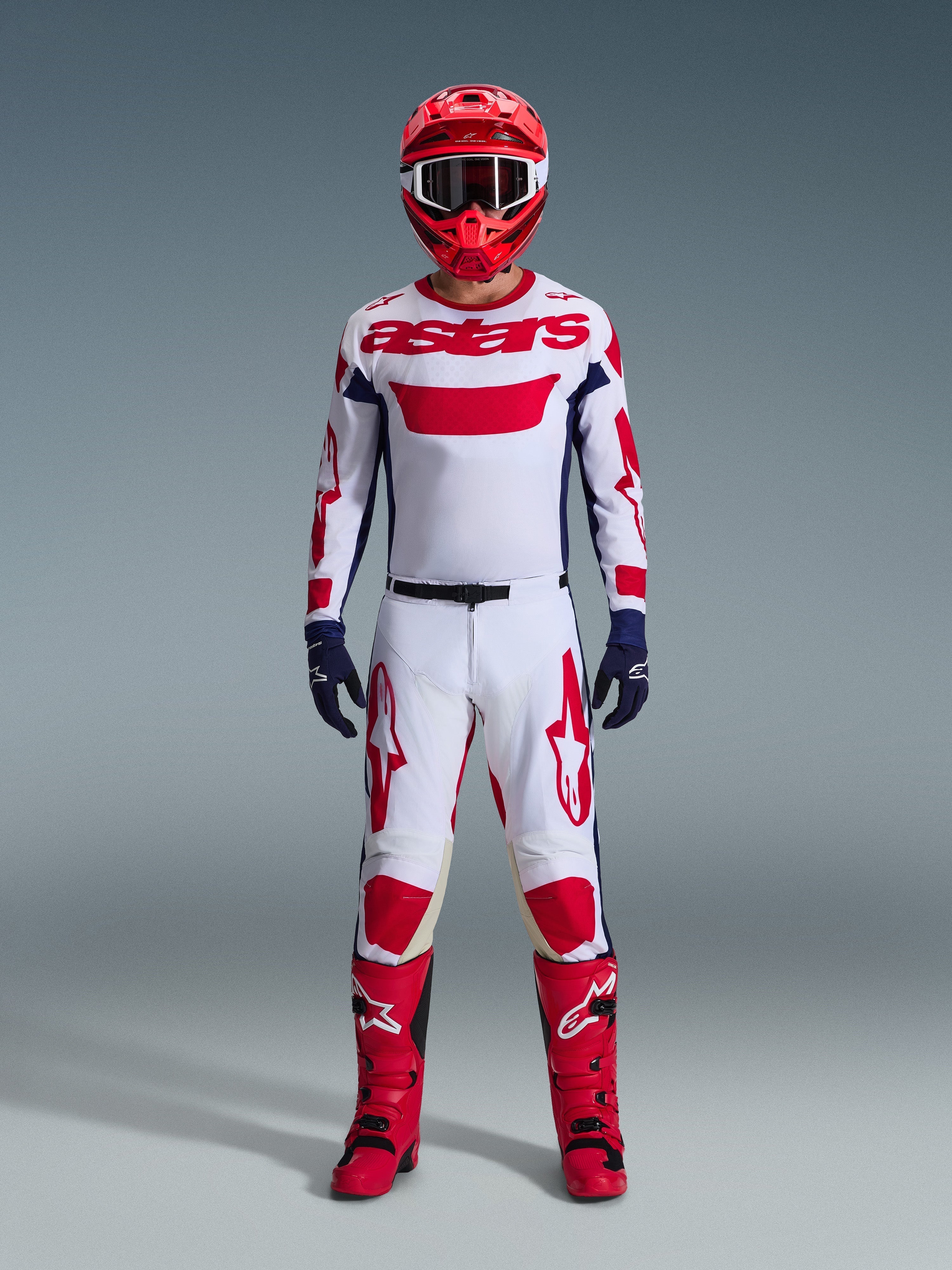 Pilota di motocross che indossa il completo Alpinestars Racer Riway 2026, maglia bianca con marchio "astars" rosso e pannelli laterali blu, pantaloni bianchi con grafiche rosse, casco da gara e stivali rossi, guanti blu scuro, abbigliamento completo coordinato da motocross