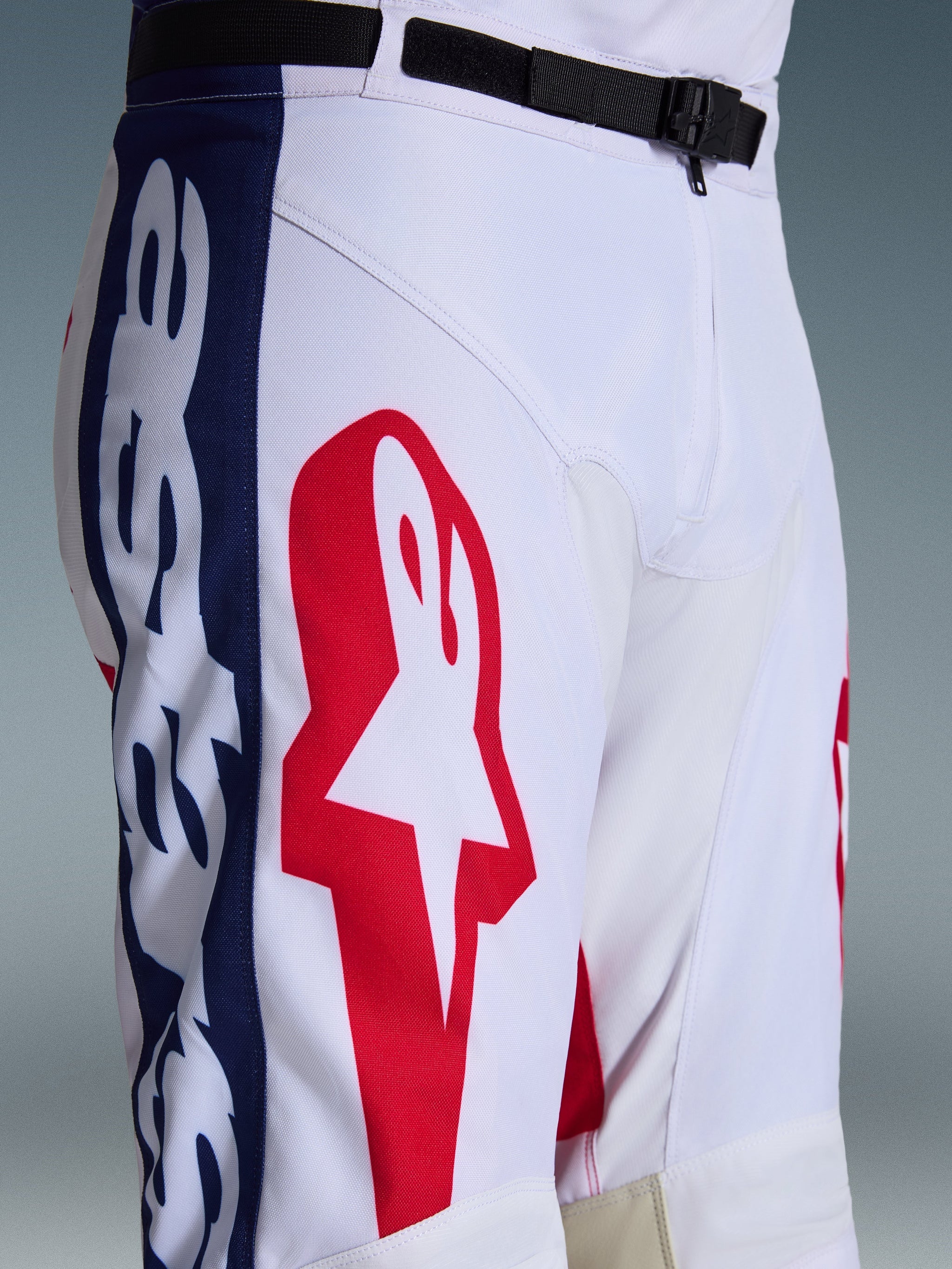 Pantaloni Alpinestars Racer Riway 2026, pantaloni da motocross, bianco blu rosso, primo piano della parte superiore della gamba e della vita che mostra il grande logo a stella rosso su tessuto bianco, pannelli laterali blu con marchio e cintura regolabile nera in vita con fibbia magnetica SX2, design sportivo da corsa fuoristrada