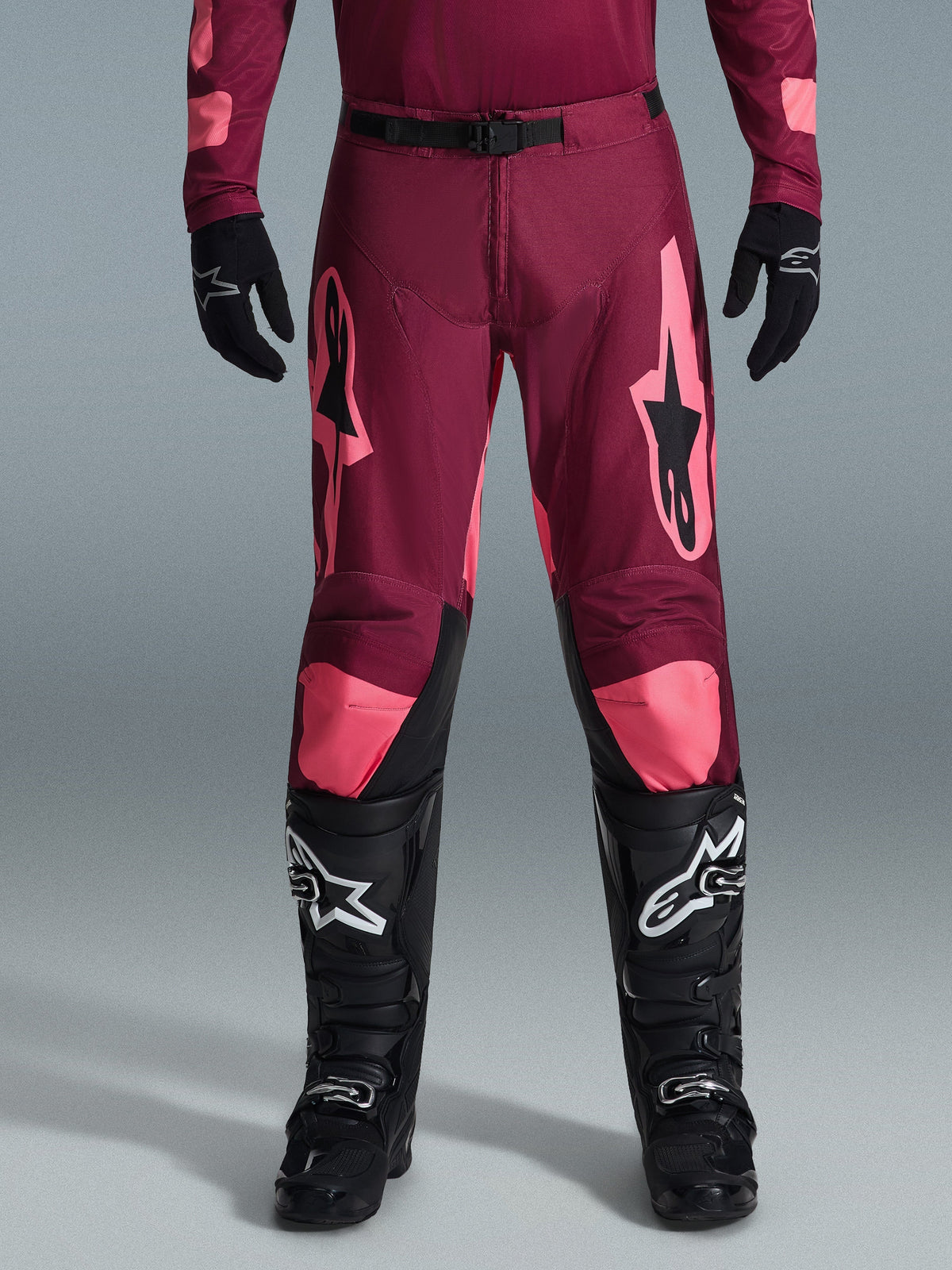 Alpinestars 2026 Racer Riway Pants, pantaloni da motocross, color corallo rosso scuro, indossati con guanti e stivali neri, con grandi loghi Alpinestars color corallo sulle cosce, fibbia nera in vita e design della gamba pre-curvato per corse fuoristrada