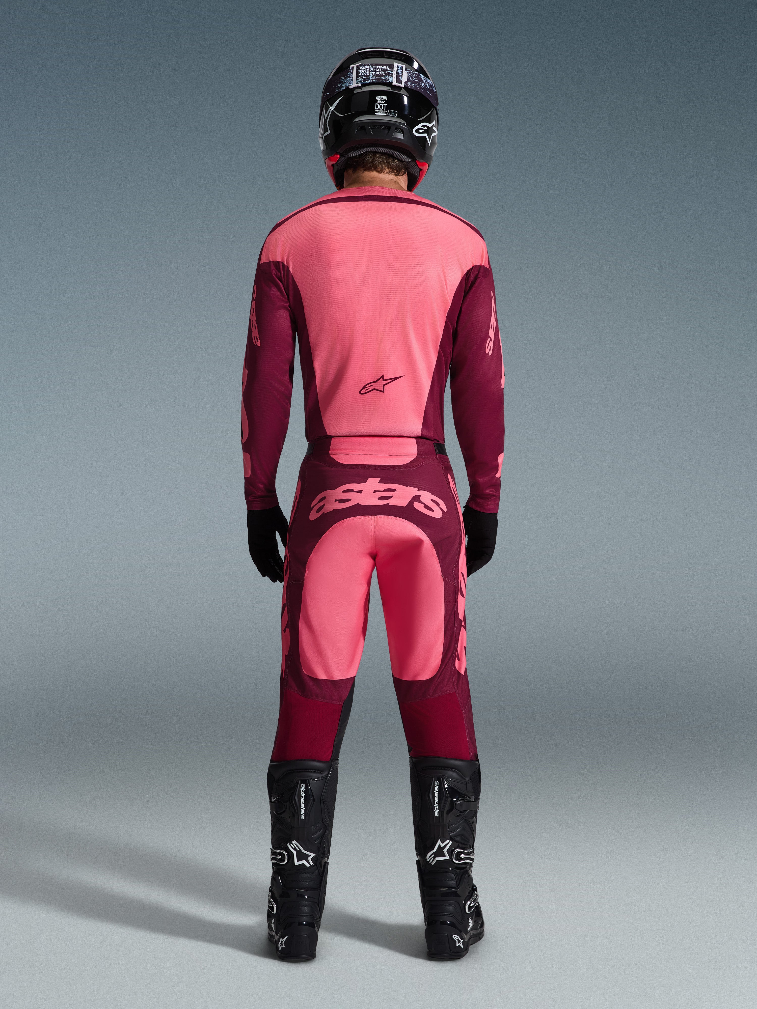 Pilota di motocross visto da dietro che indossa i pantaloni Alpinestars 2026 Racer Riway in Dark Red Coral, maglia coordinata, casco, guanti e stivali neri, con logo 'astars' sulla seduta e design bicolore rosso.
