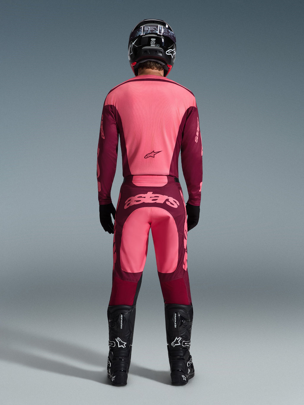Vista posteriore di un pilota di motocross con completo Alpinestars Racer Riway 2026, maglia rosa e bordeaux, pantaloni bordeaux con logo 'astars' rosa chiaro sulla seduta, casco, stivali e guanti neri, outfit coordinato per prestazioni off-road