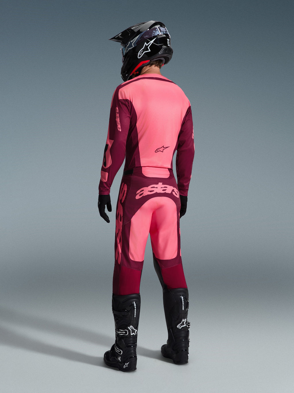 Pantaloni Alpinestars Racer Riway 2026, pantaloni da motocross, Dark Red Coral, vista posteriore di un pilota che indossa maglia coordinata, casco nero e stivali neri, con vistoso branding corallo sulla seduta e vestibilità atletica ergonomica per gare off-road