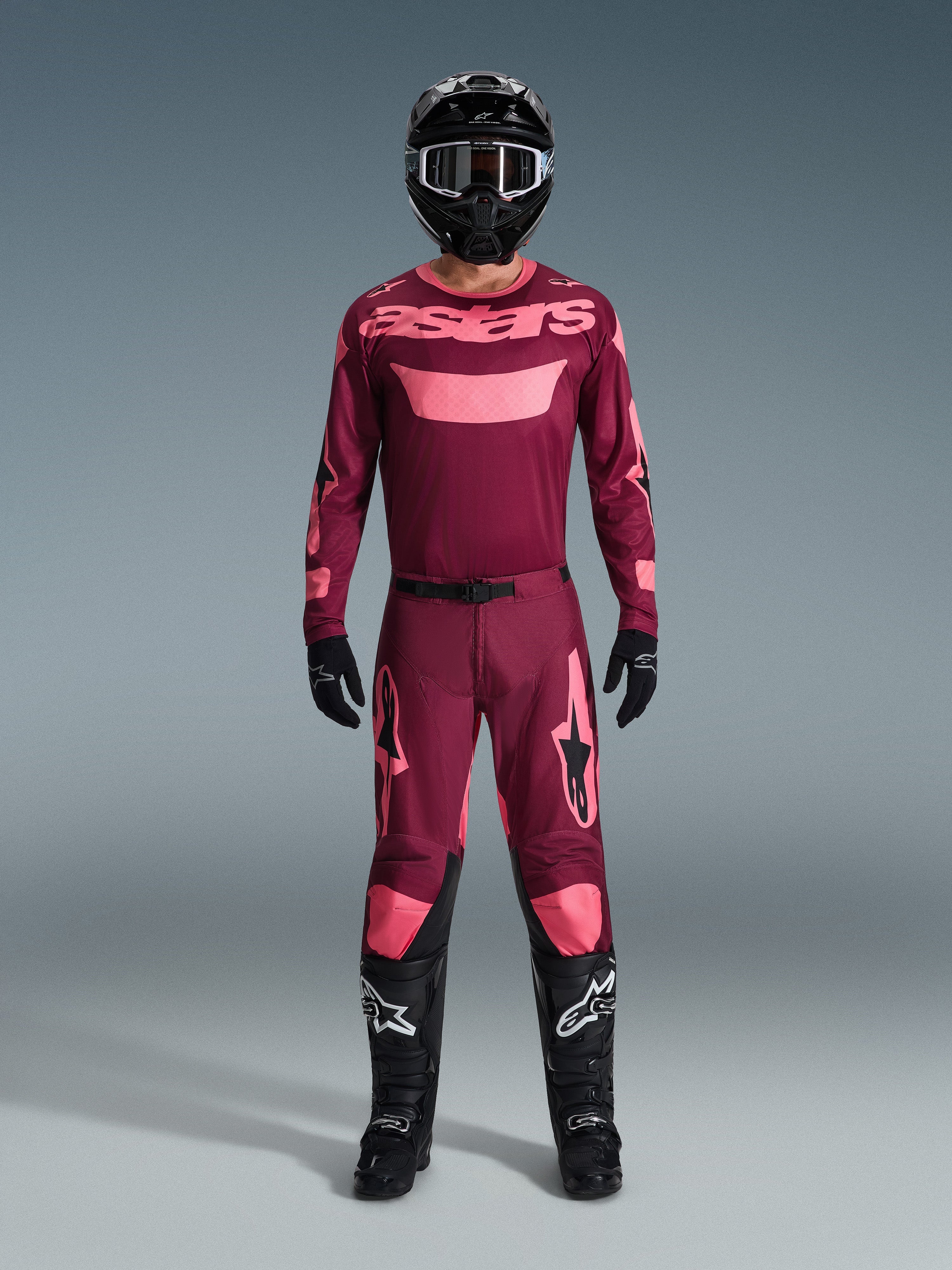 Pilota di motocross che indossa il completo Alpinestars 2026 Racer Riway, maglia bordeaux con logo 'astars' rosa, pantaloni bordeaux coordinati con accenti rosa su ginocchia e gambe, casco nero, guanti neri e stivali neri, completo tecnico per gare off-road.