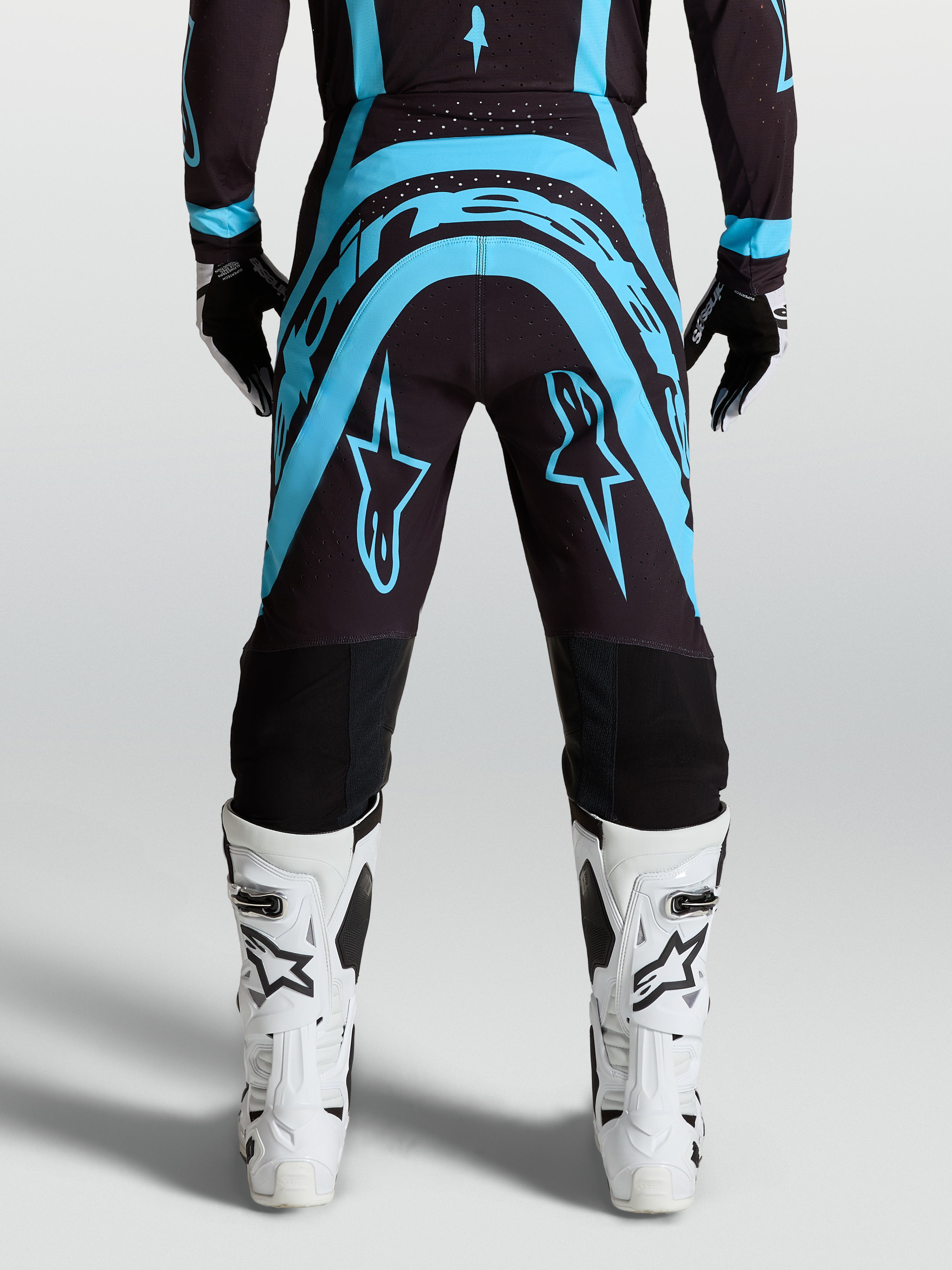 Alpinestars Pantaloni Supertech Vesta, pantaloni da motocross nero e turchese, vestibilità pro-level race-fit con stivali da moto bianchi, abbigliamento fuoristrada tecnico.