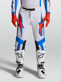 Alpinestars Pantaloni Supertech Vesta, pantaloni da motocross pro-level da uomo, bianchi con pannelli blu e rossi, vestibilità atletica ed elasticizzata con perforazioni laser, indossati da un pilota con stivali bianchi.