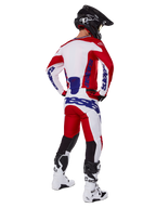 Pantaloni Alpinestars Racer Veil 2025, pantaloni da motocross, rosso e bianco con accenti blu, visti da dietro su un pilota con attrezzatura completa, logo Alpinestars audace, pannelli traspiranti sulla parte inferiore delle gambe, stivali bianchi e casco nero coordinati.