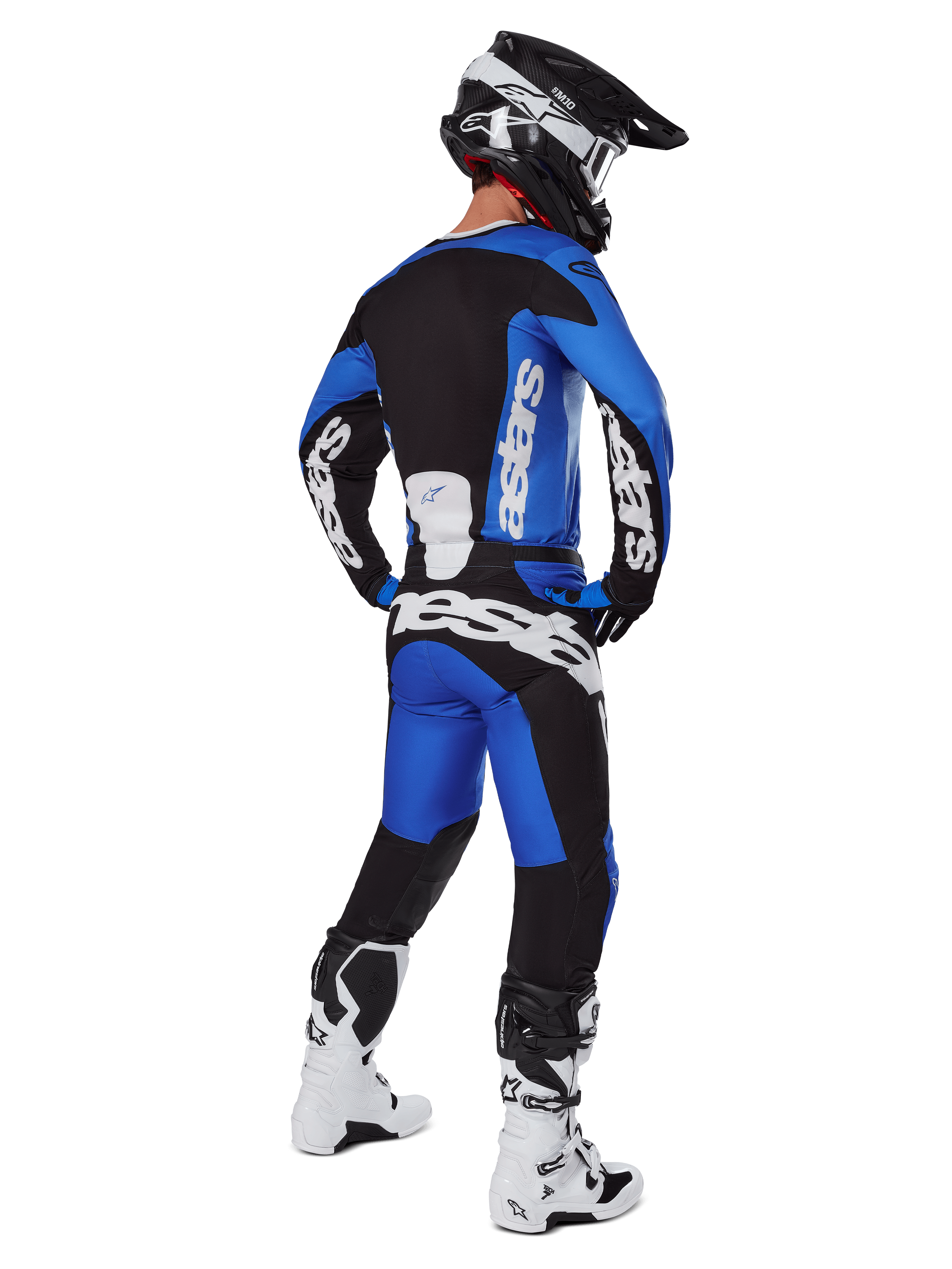 Pilota di motocross visto da dietro che indossa pantaloni Alpinestars Racer Veil 2025 blu e neri, maglia a maniche lunghe coordinata con logo bianco, stivali Tech 7 bianchi e neri e casco SM10 nero, set completo di abbigliamento per corse fuoristrada