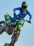 Pilota di motocross a metà salto su una moto verde, che indossa pantaloni Alpinestars 2025 Racer Veil in blu e nero, maglia blu coordinata con logo bianco sul petto, stivali bianchi e neri e casco nero, abbigliamento off-road ad alte prestazioni per gare.