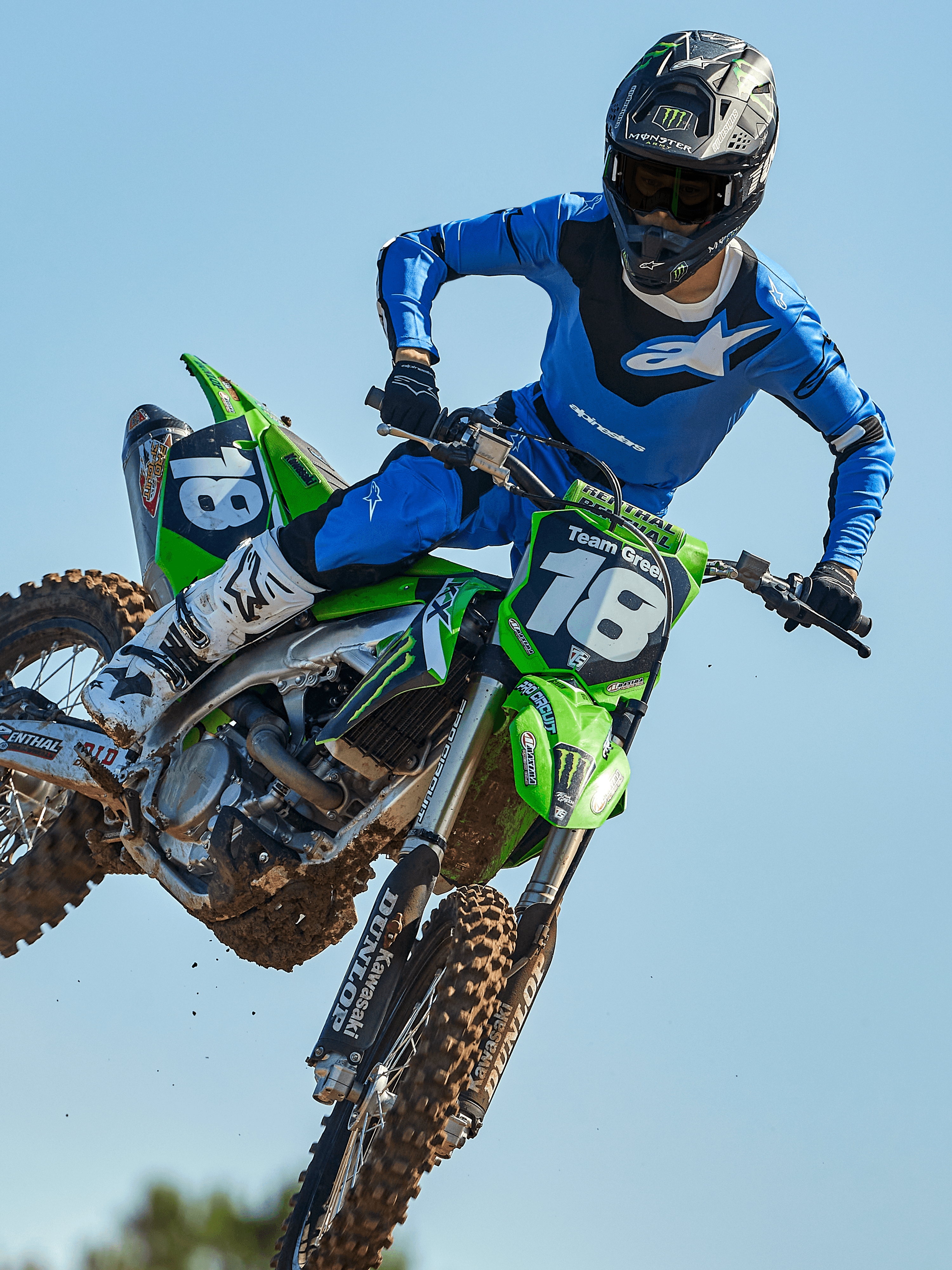 Pilota di motocross a metà salto su una moto verde, che indossa pantaloni Alpinestars 2025 Racer Veil in blu e nero, maglia blu coordinata con logo bianco sul petto, stivali bianchi e neri e casco nero, abbigliamento off-road ad alte prestazioni per gare.