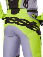 Alpinestars Pantaloni Racer Veil 2025, pantaloni da motocross, grigio e giallo fluo, vista posteriore con grande logo nero sulla seduta, indossati con maglia e guanti coordinati, design tecnico per fuoristrada