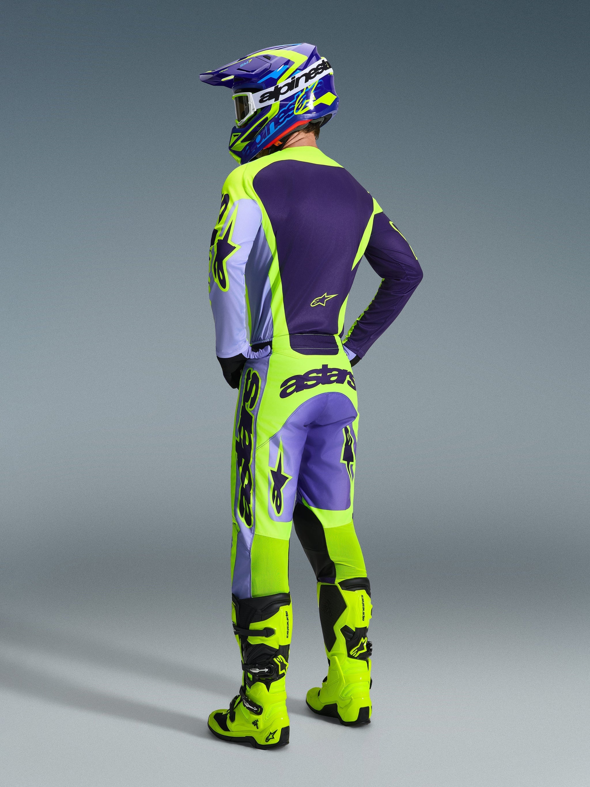 Pilota di motocross visto da dietro con indosso i pantaloni Alpinestars 2026 Racer Portl in Purple Yellow Fluo, abbinati a maglia e casco coordinati in viola e giallo e stivali da gara giallo vivace, con l'evidente marchio 'astars' sulla seduta e i loghi a stella sulle gambe.