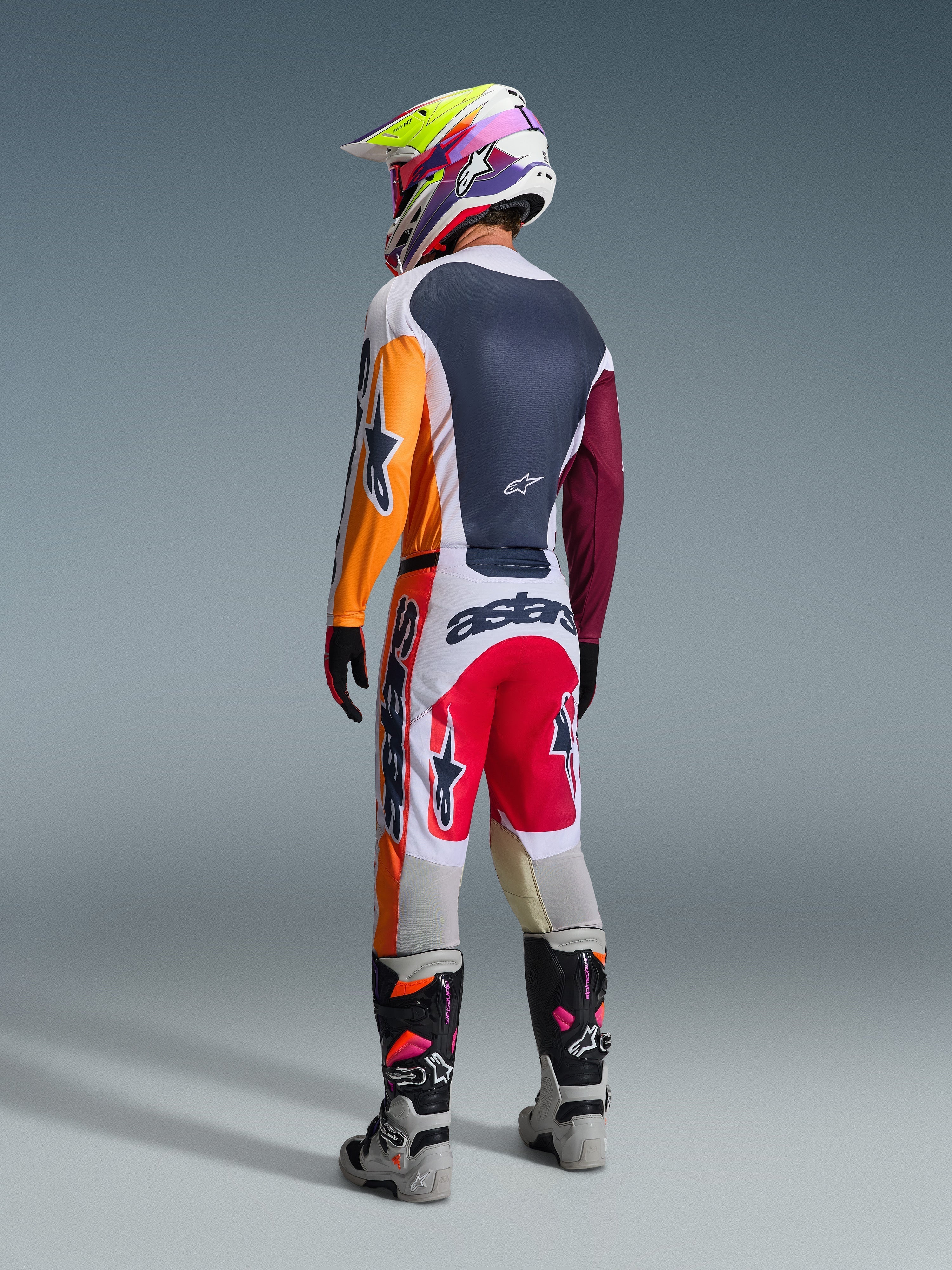 Pilota di motocross visto da dietro con abbigliamento completo da gara, pantaloni Alpinestars 2026 Racer Portl in rosso, grigio chiaro e grigio scuro, maglia coordinata con maniche arancioni e bordeaux, casco con grafica multicolore, stivali da fuoristrada grigi e neri con dettagli rosa, completo professionale da motocross