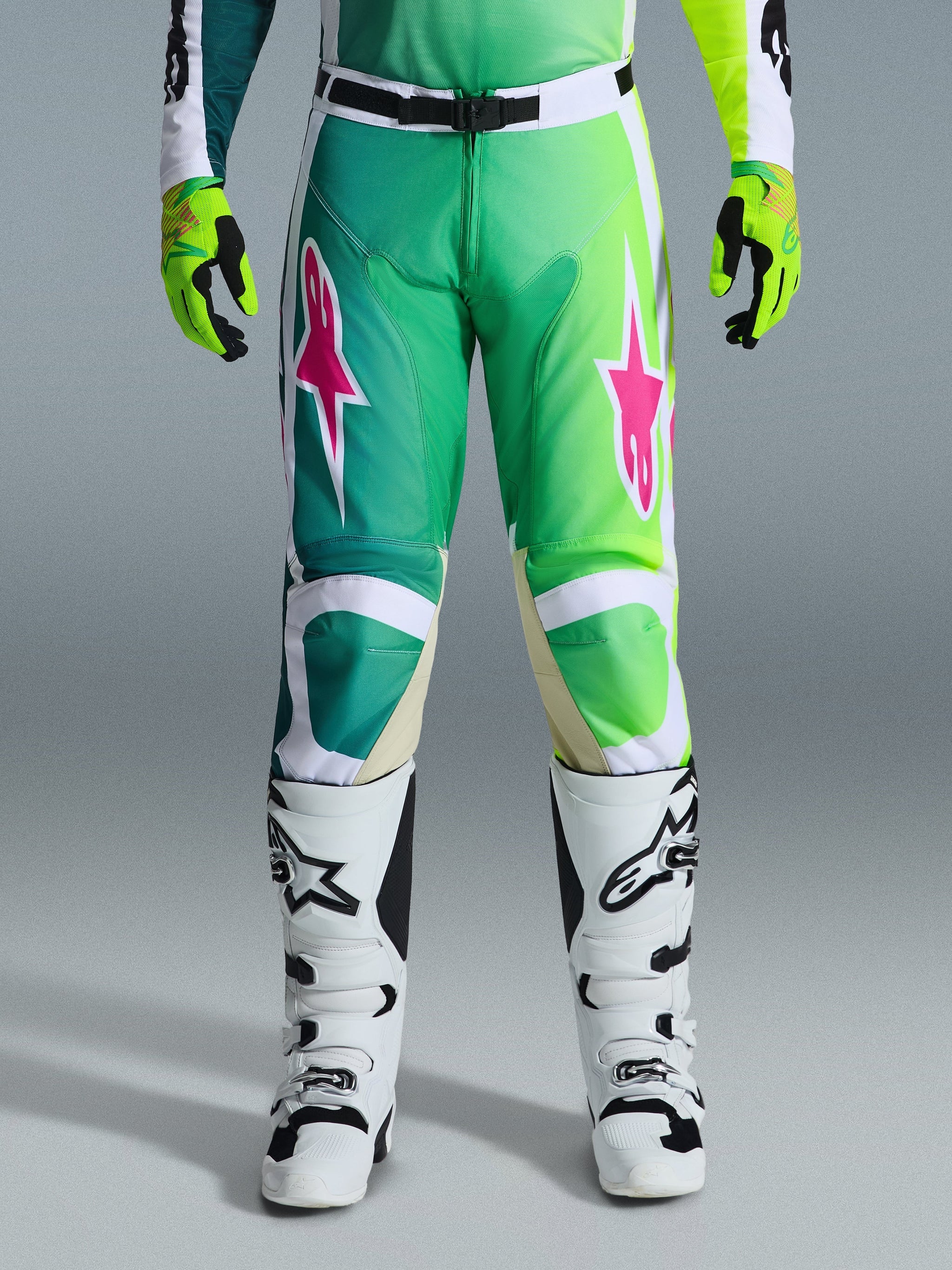 Alpinestars 2026 Racer Portl Pants, pantaloni da motocross, verde, bianco e nero con loghi a stella rosa, indossati con stivali bianchi e neri e guanti verde lime, caratterizzati da un design sfumato e vestibilità ergonomica per gare off-road