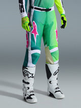 Alpinestars Racer Portl Pants 2026, pantaloni da motocross, verde, bianco e nero con loghi rosa, indossati con stivali bianchi e guanti giallo neon, caratterizzati da grafiche audaci e fibbia regolabile nera in vita, abbigliamento tecnico per fuoristrada.