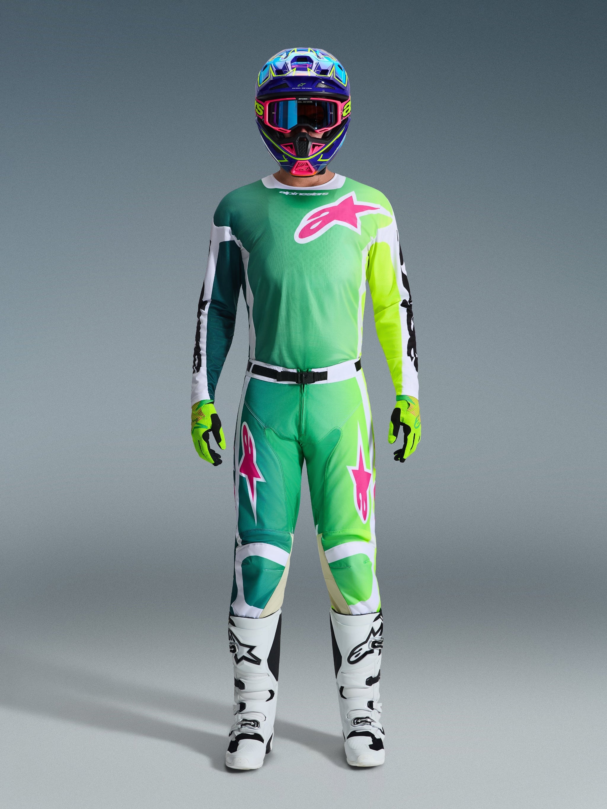 Alpinestars 2026 Racer Portl Pants, pantaloni da motocross, verde, bianco e nero con accenti rosa nel logo, indossati come parte di un set da gara completo con maglia coordinata, stivali bianchi e casco multicolore, outfit coordinato per competizioni off-road professionali.