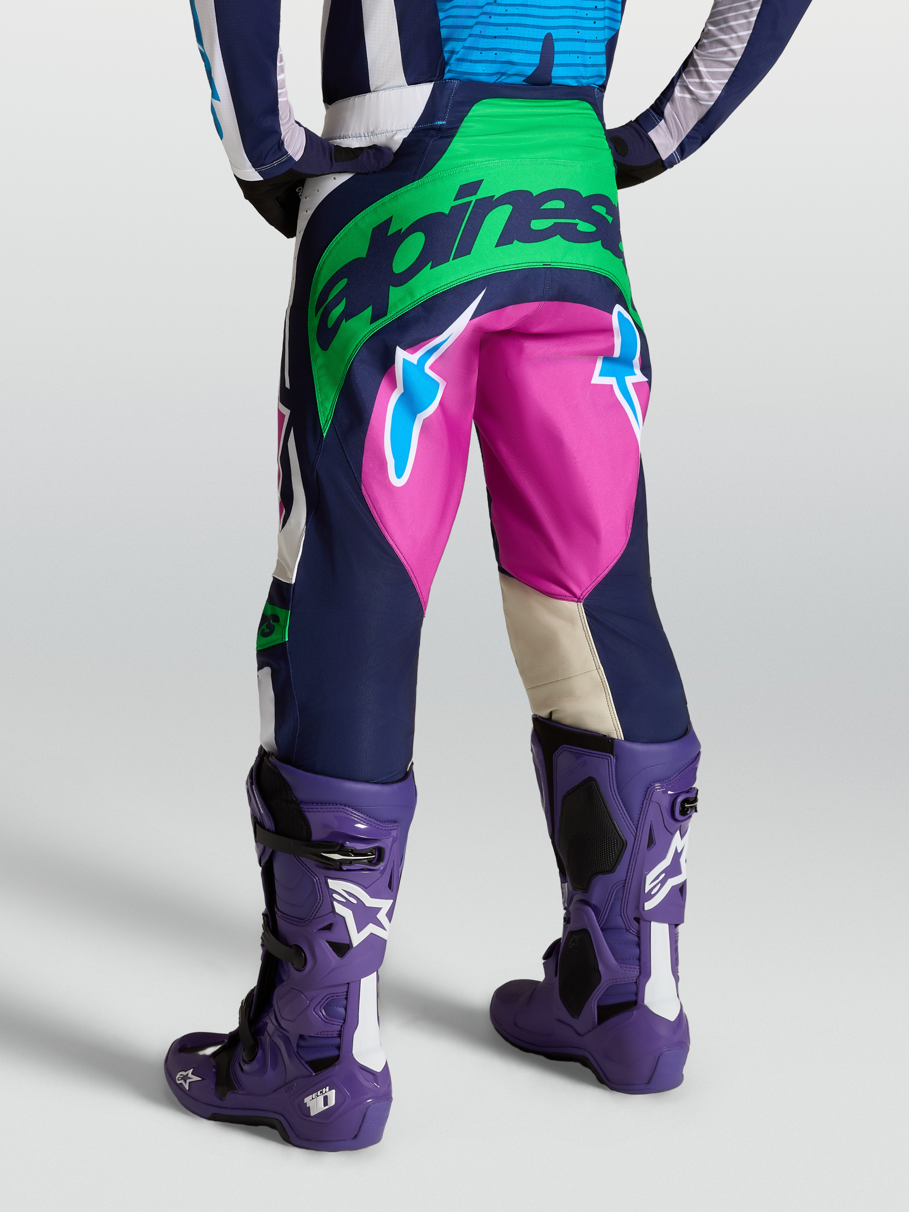 Alpinestars Pantaloni Techstar Maven, pantaloni motocross da uomo, blu e verde con dettagli fucsia, vista posteriore su modello con stivali Tech 10 viola.