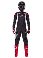 Alpinestars 2025 Fluid Haul Gear Set, pilota di motocross in abbigliamento completo, maglia nera con dettagli rossi sulle spalle e loghi bianchi, pantaloni neri con logo a stella bianco, stivali rosso acceso, guanti rossi e neri, casco nero con occhiali bianchi, look coordinato per prestazioni off-road