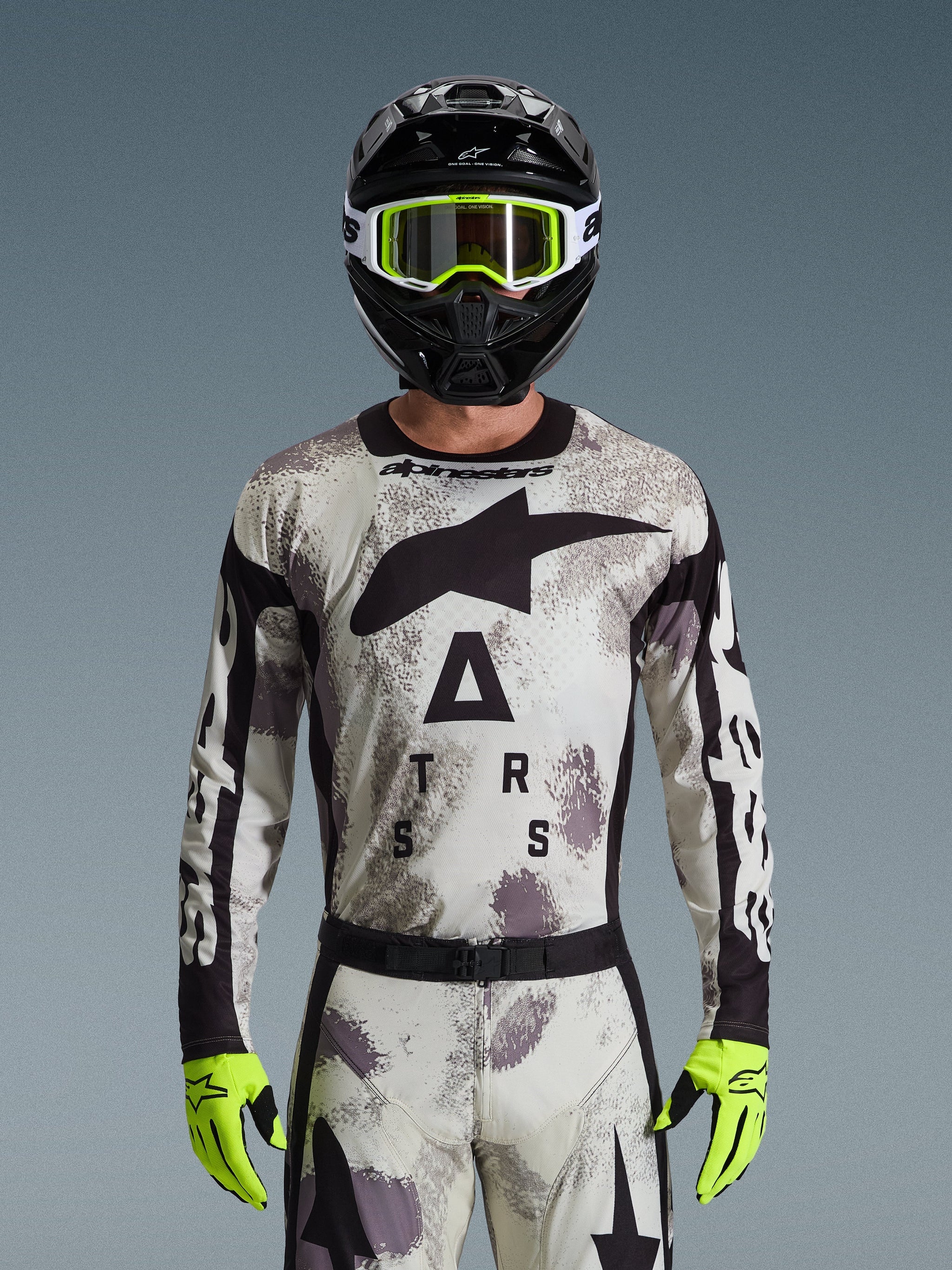 Pilota di motocross che indossa la maglia Alpinestars Racer Lahnd 2026, motivo Sand Camo in grigio e bianco, grande logo Astar nero e branding sul petto, abbinata a un casco nero, occhiali e guanti giallo neon, abbigliamento tecnico per il fuoristrada