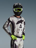 Maglia Alpinestars Racer Lahnd 2026, maglia da motocross, Sand Camo con motivo grigio e nero, grande logo Astars sul petto, indossata con casco nero, maschera e guanti giallo neon, abbigliamento tecnico per corse fuoristrada