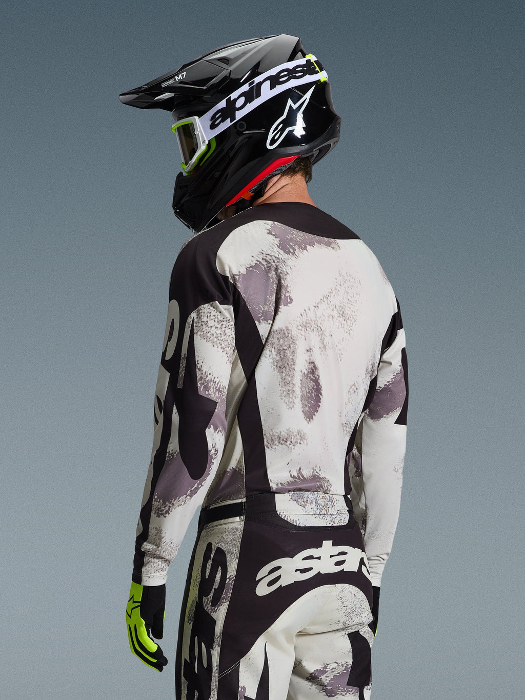 Pilota da motocross visto di spalle che indossa il completo Alpinestars 2026 Racer Lahnd, maglia e pantaloni Sand Camo grigio e bianco con dettagli neri, casco nero con logo Alpinestars bianco, guanti giallo neon e neri, abbigliamento tecnico coordinato da fuoristrada