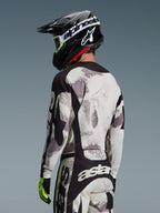 Pilota da motocross visto di spalle che indossa il completo Alpinestars 2026 Racer Lahnd, maglia e pantaloni Sand Camo grigio e bianco con dettagli neri, casco nero con logo Alpinestars bianco, guanti giallo neon e neri, abbigliamento tecnico coordinato da fuoristrada