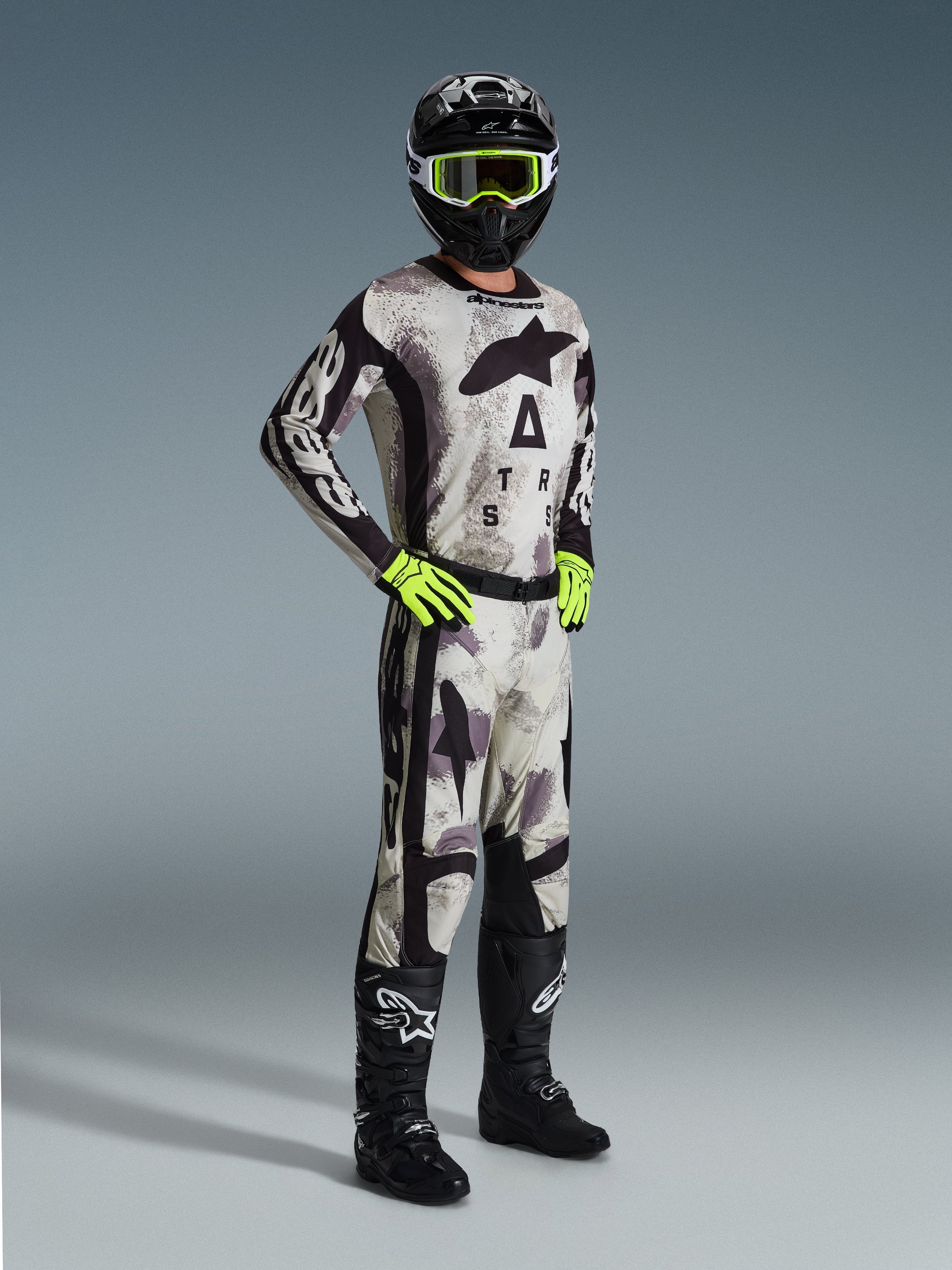 Pilota di motocross con Maglia Racer Lahnd 2026 Alpinestars in Sand Camo, motivo mimetico grigio e nero, pantaloni da motocross coordinati, guanti giallo neon, casco nero con occhiali gialli e stivali neri, abbigliamento completo professionale da gara fuoristrada