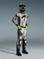Alpinestars 2026 Racer Lahnd Gear Set, pilota di motocross con equipaggiamento completo da gara in Sand Camo e grigio, maglia e pantaloni coordinati con dettagli neri e loghi evidenti, maschera e guanti giallo neon, casco nero e stivali neri.