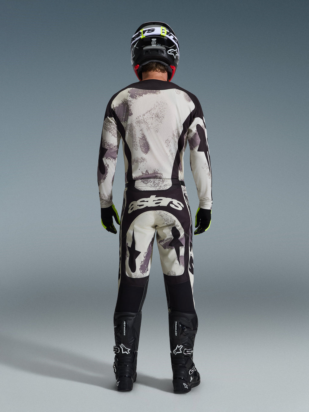 Pilota di motocross visto da dietro con pantaloni Alpinestars 2026 Racer Lahnd in Sand Camo, maglia a maniche lunghe coordinata con motivo camouflage grigio, stivali da motocross neri, casco nero e logo "astars" sulla seduta, progettati per le competizioni off-road professionali