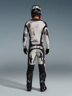 Pilota di motocross con completo Alpinestars Racer Lahnd 2026, vista posteriore, maglia e pantaloni Sand Camo con motivo astratto grigio chiaro e nero, casco nero, guanti nero e giallo neon, stivali neri, marchio 'astars' sul retro dei pantaloni