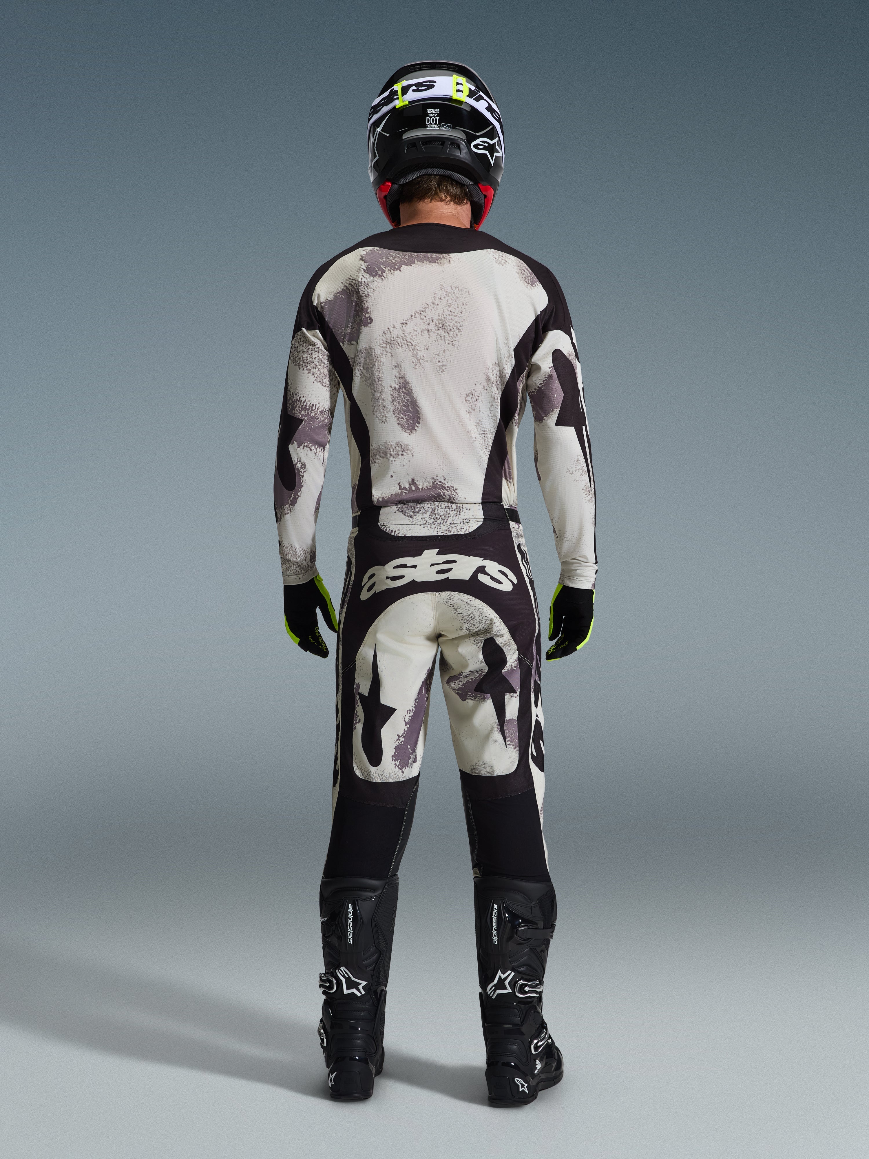 Pilota di motocross visto da dietro con maglia 2026 Racer Lahnd e pantaloni coordinati in Sand Camo, caratterizzati da un motivo astratto crema e grigio con pannelli neri, logo 'astars' sui pantaloni, stivali Alpinestars neri e casco nero per un look da gara off-road completo.