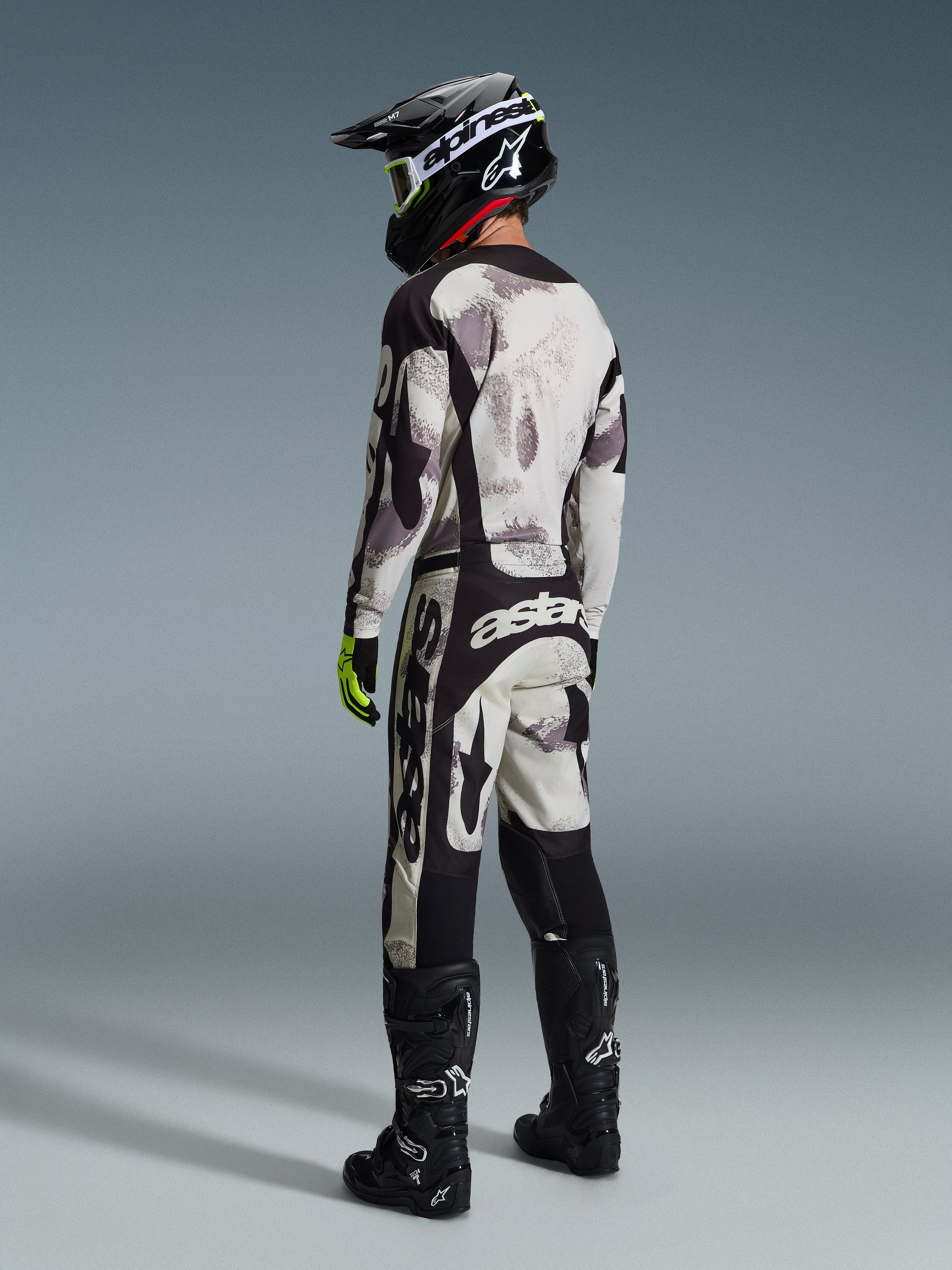 Pilota di motocross con abbigliamento da gara completo, Maglia Racer Lahnd 2026 e pantaloni coordinati in Sand Camo, caratterizzati da un motivo mimetico grigio e nero con evidenti loghi Alpinestars, casco nero, stivali neri e guanti giallo neon, vista posteriore di profilo.