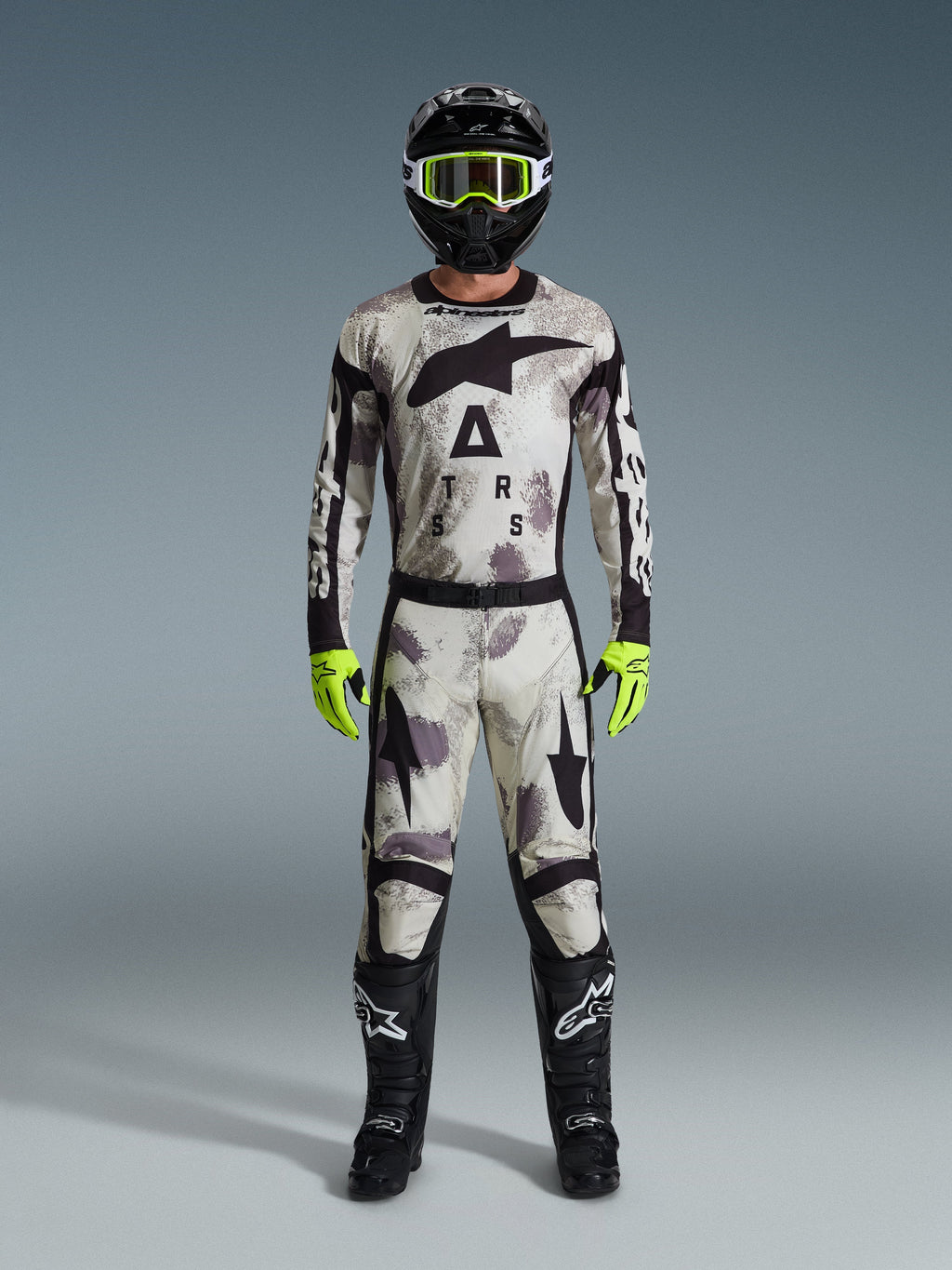 Pilota di motocross con abbigliamento da gara completo, pantaloni Alpinestars 2026 Racer Lahnd e maglia coordinata in grigio Sand Camo, casco nero con occhiali giallo neon, guanti giallo neon e stivali neri, outfit professionale coordinato per corse off-road